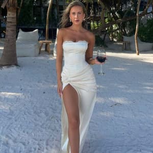 Woman in a white strapless dress standing on a sandy beach holding a glass of red wine.
