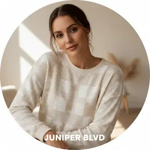 Woman wearing a white sweater with 'Juniper Blvd' branding in a blurred indoor setting.