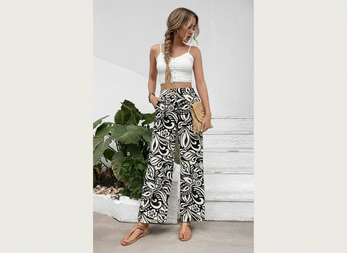 Woman wearing a white top and patterned pants standing against a light-colored wall with plants.