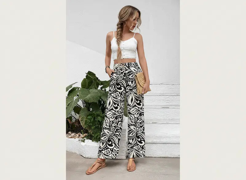 Woman wearing a white top and patterned pants standing against a light gray wall with a plant in the background.