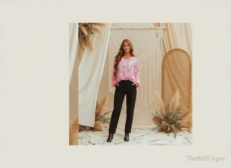 Woman wearing a pink floral top and black pants standing in a decorative room with plants and arches.