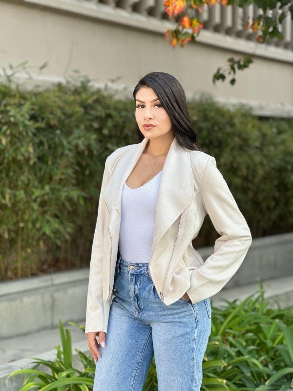 Woman wearing a white jacket and blue jeans standing outdoors with greenery in the background.
