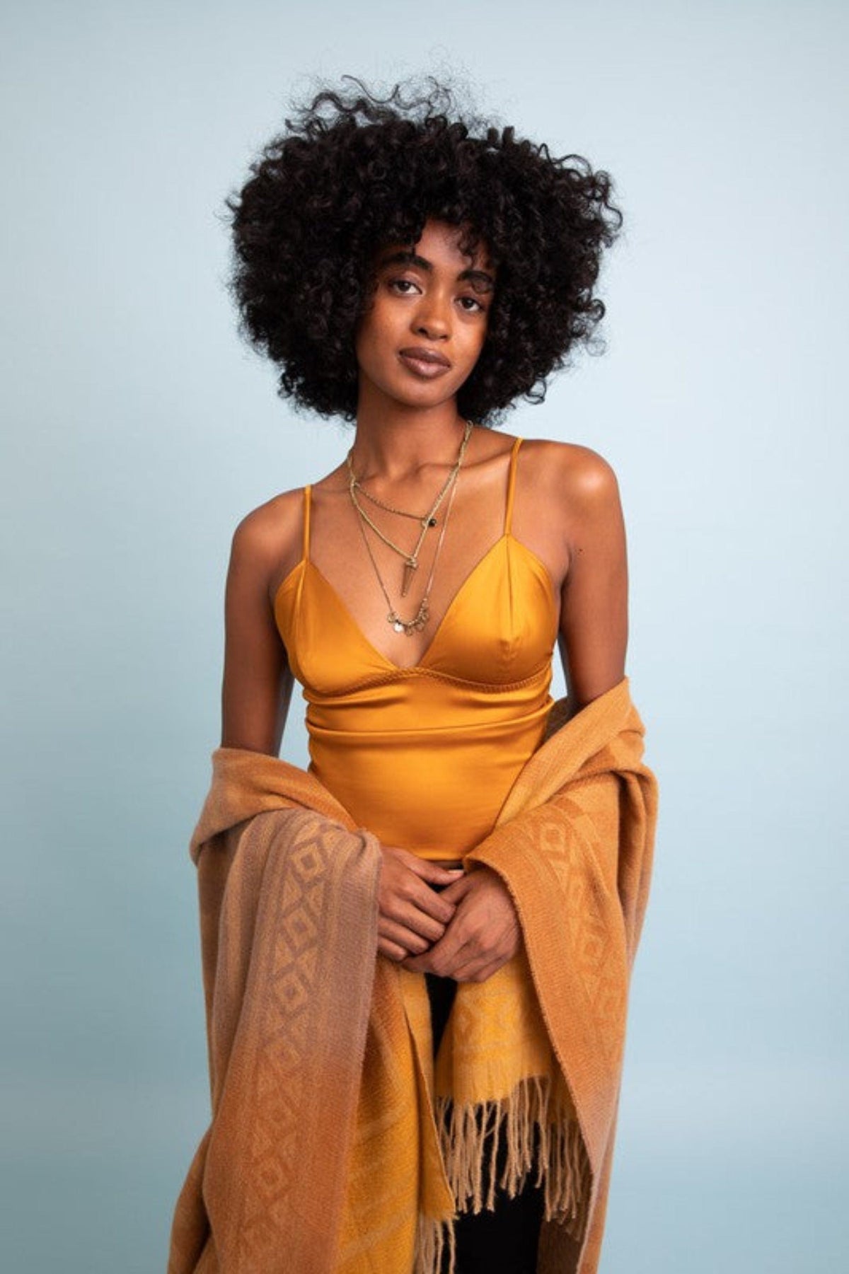 Woman casually posing in a low-cut gold tank top with a shrug wrapped around her arms, showcasing the trendy styles from our Festival Vibes collection.