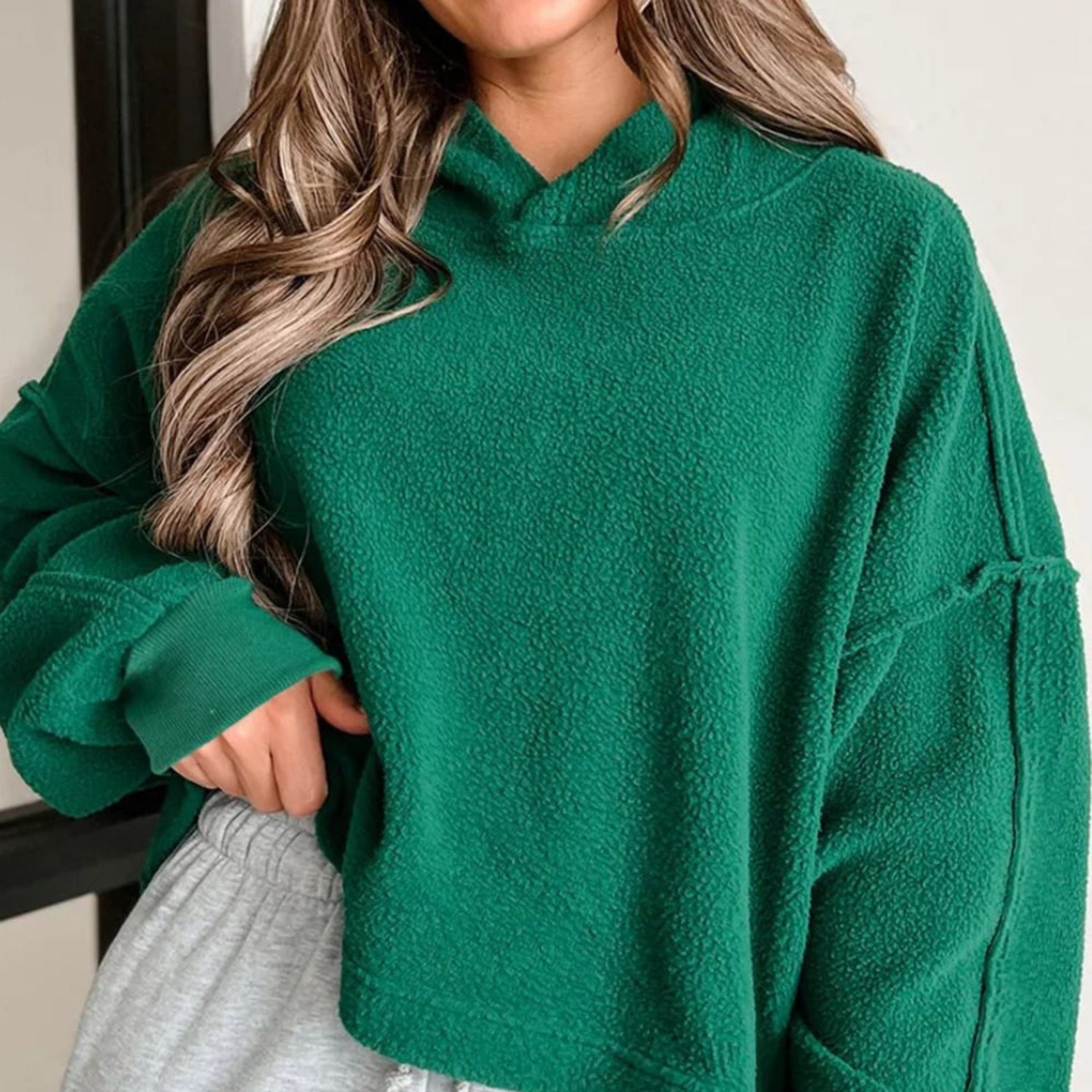 A model wearing a women's oversized comfortable-inspired hoodie from the hoodie and sweatshirt collection at the802Gypsy, showcasing green fabric with a hooded neckline and a longer cropped style with a simple backdrop where the model is near a wall.